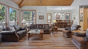 A spacious living room with wooden flooring, leather sofas, a dining area with wooden furniture, and large windows offering a view of trees.