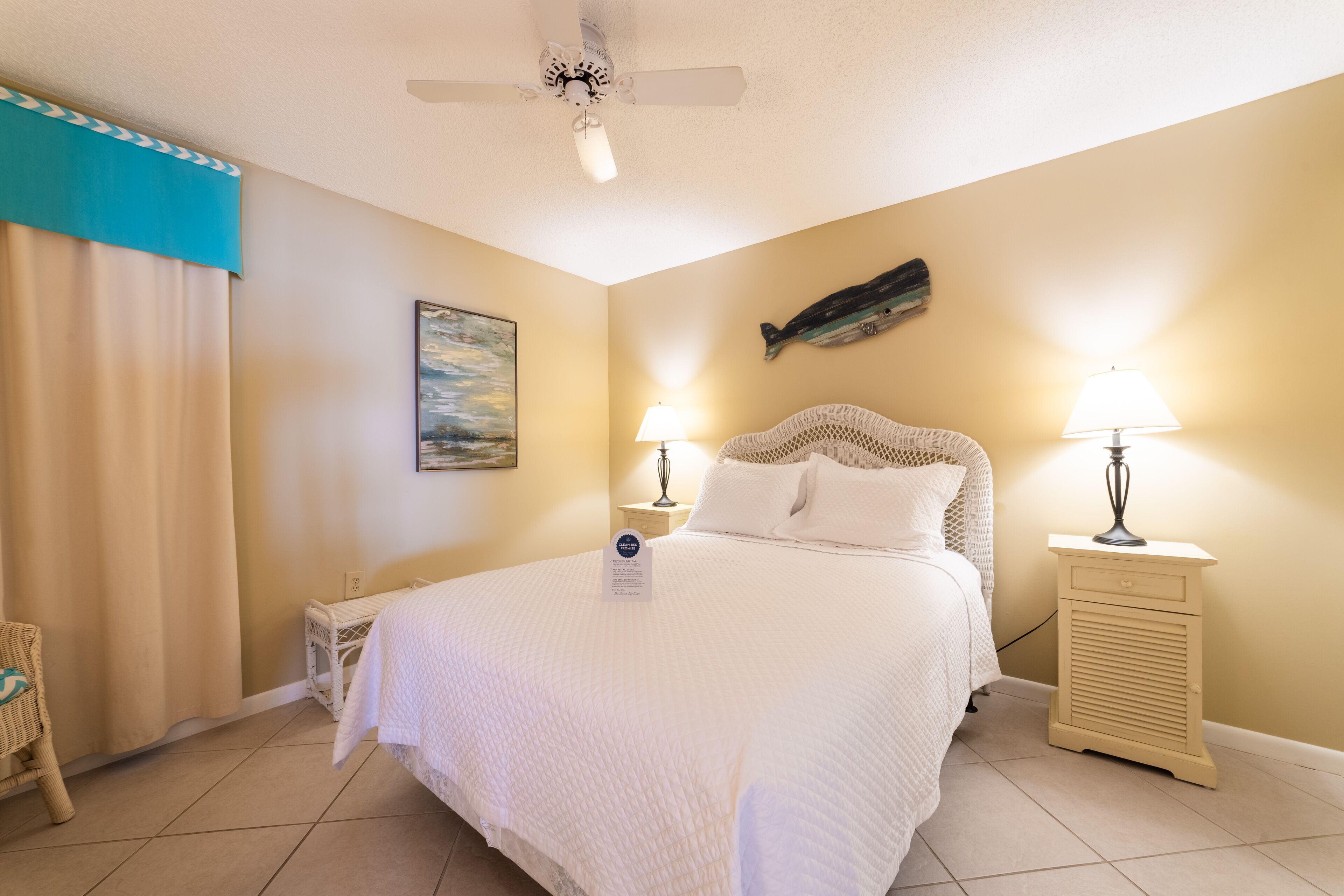 A bedroom with a bed, two bedside lamps, a ceiling fan, a fish decoration, a painting, and a small table with a lamp.