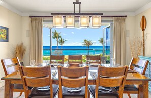 A dining area with a table set for four, overlooking a beach with palm trees and the ocean. - Maui Resort Rentals Dream Collection: Honua Kai Hokulani 202 – Direct Oceanfront 3BR w/ BBQ on Wraparound Balcony (Lahaina)