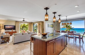 Relax in a spacious living area with a sofa set, dining table, and kitchen island, offering a view of the ocean and palm trees.