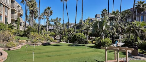 A golf course with a well-maintained green, surrounded by palm trees and a hotel building.