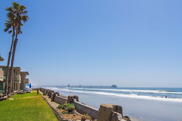 A beach with a pier extending into the ocean, a grassy area with palm trees, and a building with balconies.