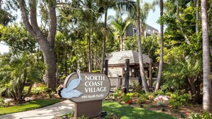 Un panneau indiquant l’entrée de North Coast Village, situé au 999 North Pacific, entouré d’une végétation luxuriante et bordé d’un sentier.