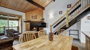 Un salon chaleureux avec une table à manger en bois, un canapé en cuir et une cheminée. Un escalier avec une rampe en bois mène à l’étage supérieur.