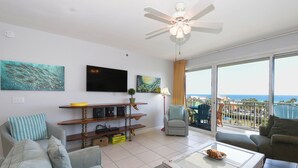 Ein geräumiges Wohnzimmer mit großem Fenster, Deckenventilator, Flachbildfernseher und einem Balkon mit Meerblick.