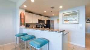 A kitchen with white cabinets, a granite countertop, and a stainless steel refrigerator. There are three blue bar stools and a surfboard decoration on the wall. - Bright 4th Floor Condo w/ Gulf Views & Modern Touches on Scenic 98 in Destin! 🏖 (Destin)