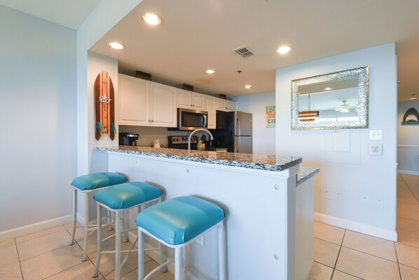 A kitchen with white cabinets, a granite countertop, and a stainless steel refrigerator. There are three blue bar stools and a surfboard decoration on the wall. - Bright 4th Floor Condo w/ Gulf Views & Modern Touches on Scenic 98 in Destin! 🏖 (Destin)