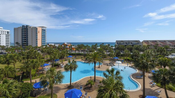 Ein Resort mit einem großen Schwimmbad, umgeben von Palmen und Liegestühlen, mit Blick auf den Ozean und Hotelgebäuden im Hintergrund.