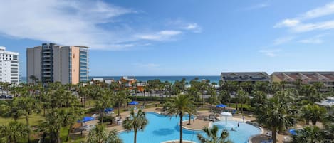 A resort with a large swimming pool surrounded by palm trees and lounge chairs, with a view of the ocean and hotel buildings in the background.