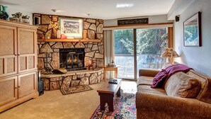 A cozy living room with a stone fireplace, a wooden cabinet, a sofa, and a large window offering a view of trees. - Atrium Bluebird Condo: Great Downtown Breck Location (Breckenridge)