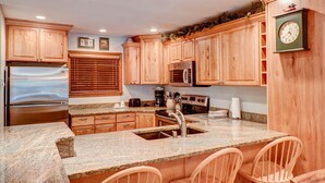 A kitchen with wooden cabinets, a stainless steel refrigerator, a microwave, an oven, granite countertops, and a clock. - Atrium Bluebird Condo: Great Downtown Breck Location (Breckenridge)
