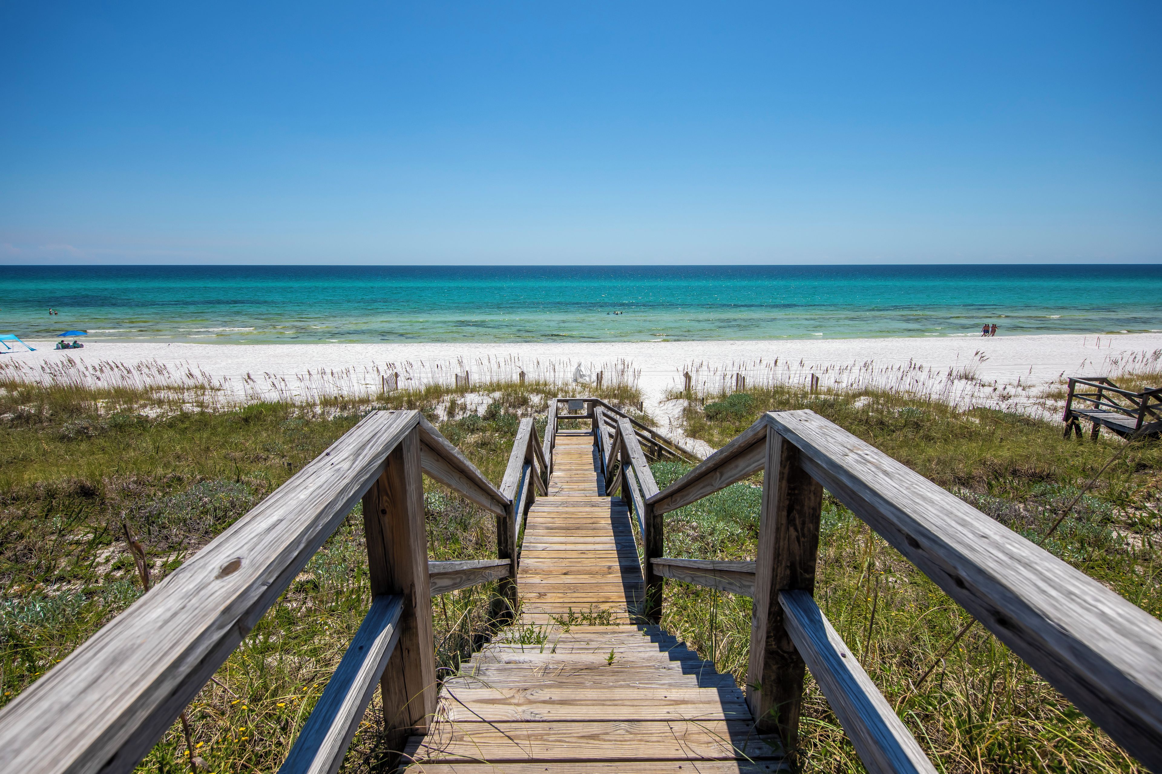 A wooden boardwalk leading to a beach with clear blue water and white sand.