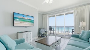 Una sala de estar con una televisión mostrando una escena de playa, un sofá, una mesa de centro, una silla y vista al océano a través de un ventanal grande.