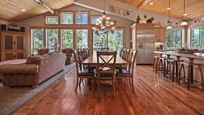 Un comedor espacioso con mesa y sillas de madera, ventanas grandes y una cocina con refrigerador y taburetes de bar.
