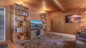 Ein gemütliches Wohnzimmer mit Holzwänden und -decke, einem großen Flachbildfernseher, Bücherregalen, einem gemusterten Teppich und einem Sofa.