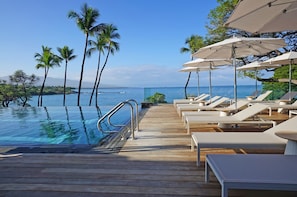 A wooden deck with a pool, lounge chairs, and umbrellas overlooking a clear blue sea.