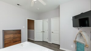 Een slaapkamer met een houten hoofdbord, een bed met witte lakens, een plafondventilator, een tv aan de muur en een inloopkast met witte deuren.