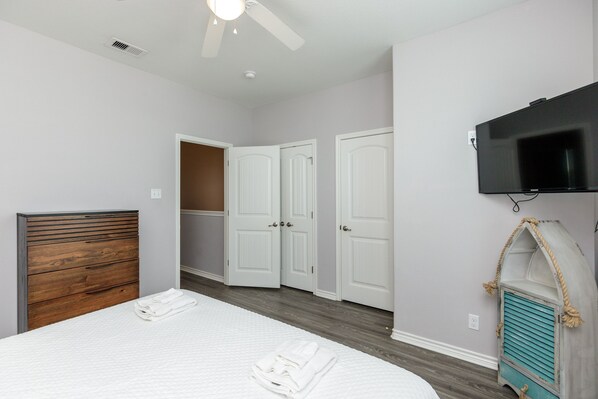 A bedroom with a wooden headboard, a bed with white linens, a ceiling fan, a TV mounted on the wall, and a closet with white doors.