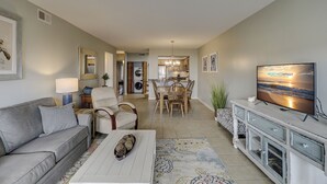 Un salon moderne avec un canapé, une table basse et un téléviseur. Il y a également un coin repas avec des chaises et une table. La pièce est décorée dans des tons neutres et possède une grande fenêtre.