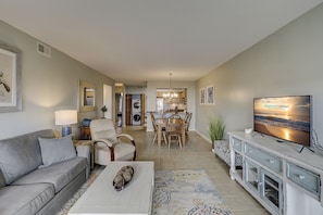 A modern living room with a sofa, coffee table, and TV. There's a dining area with chairs and a table. The room has a neutral color palette and a large window.
