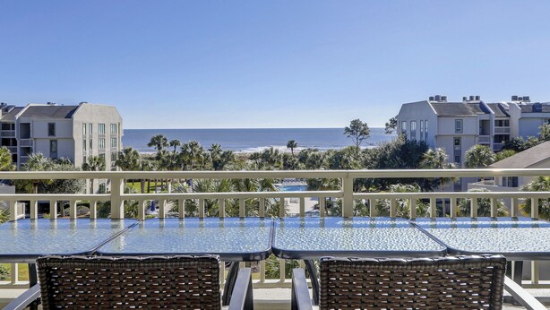 Un balcon doté de tables à plateaux en verre et de fauteuils en osier, offrant une vue sur une plage et l’océan.