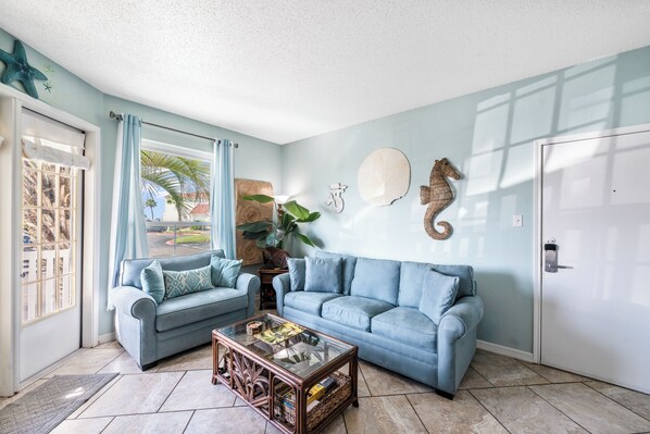 A living room with two light blue sofas, a glass coffee table, and a door with a white frame. - Walk to the Beach, Pool & Clubhouse – Pet Friendly Retreat (Corpus Christi)