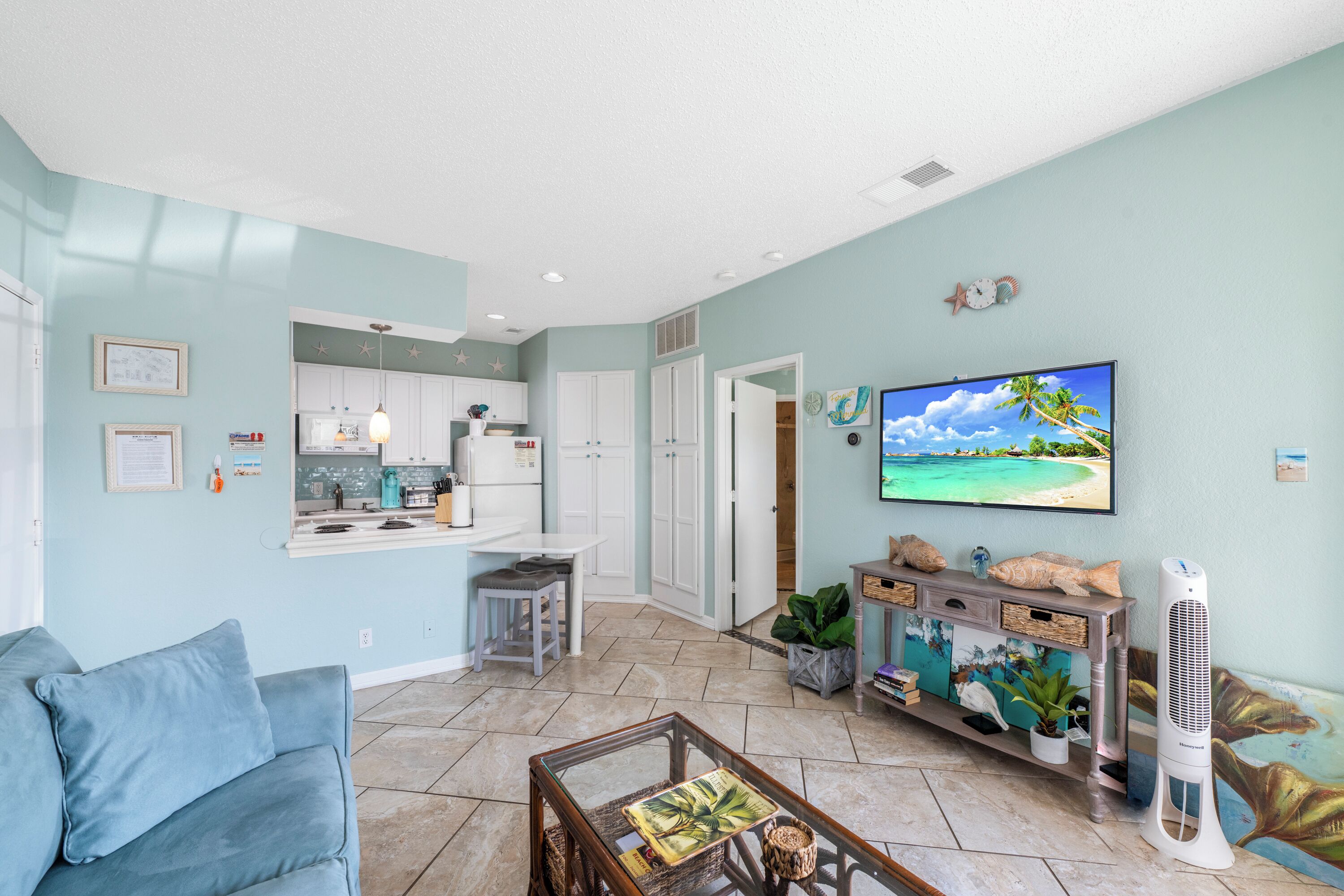 A living room with a sofa, coffee table, and a TV. There is a kitchen in the background with cabinets and a countertop.