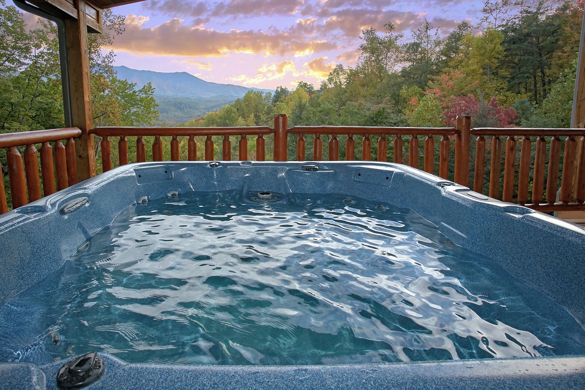 Un bain à remous doté d’une terrasse et d’une balustrade en bois, entouré d’arbres et avec une chaîne de montagnes en arrière-plan.