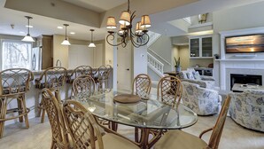 A spacious dining area with a glass-top table surrounded by wicker chairs, a chandelier, and a kitchen in the background. - 7634 Huntington-Canal views, heated pool & spa.  Quick walk to Ocean. (Hilton Head Island)
