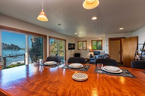 Un coin repas avec une table en bois dressée pour quatre personnes, de grandes fenêtres offrant une vue sur la mer, et un espace de détente avec un téléviseur et des fauteuils.