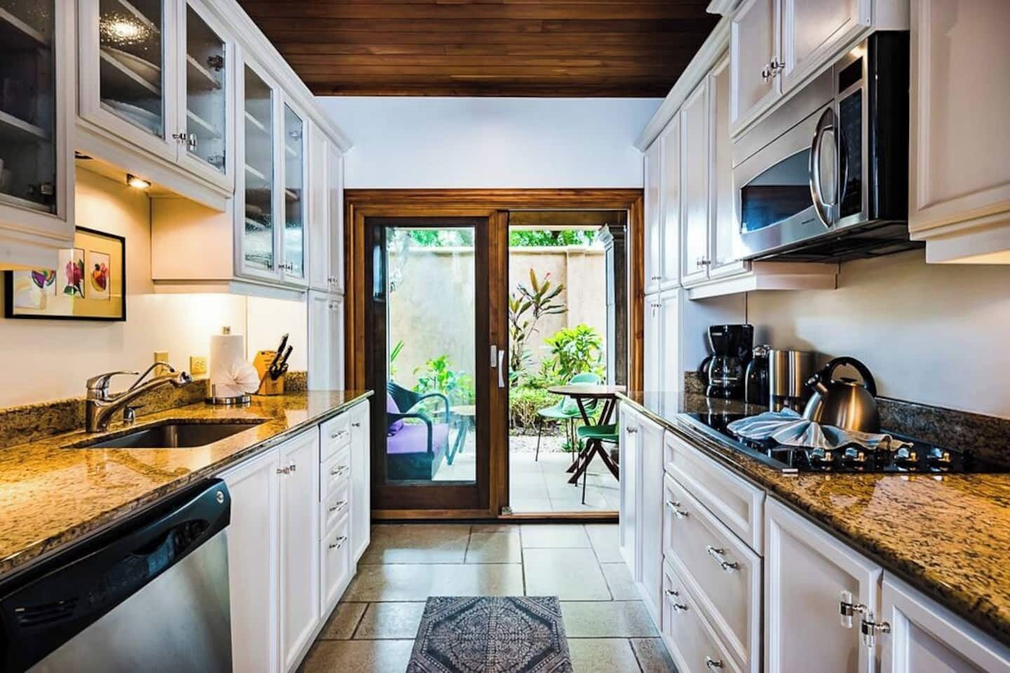 Une cuisine avec des armoires blanches, un comptoir en granit, un lave-vaisselle, une cuisinière et un four à micro-ondes. Une porte-fenêtre coulissante donne sur un patio équipé d’une table et de chaises.