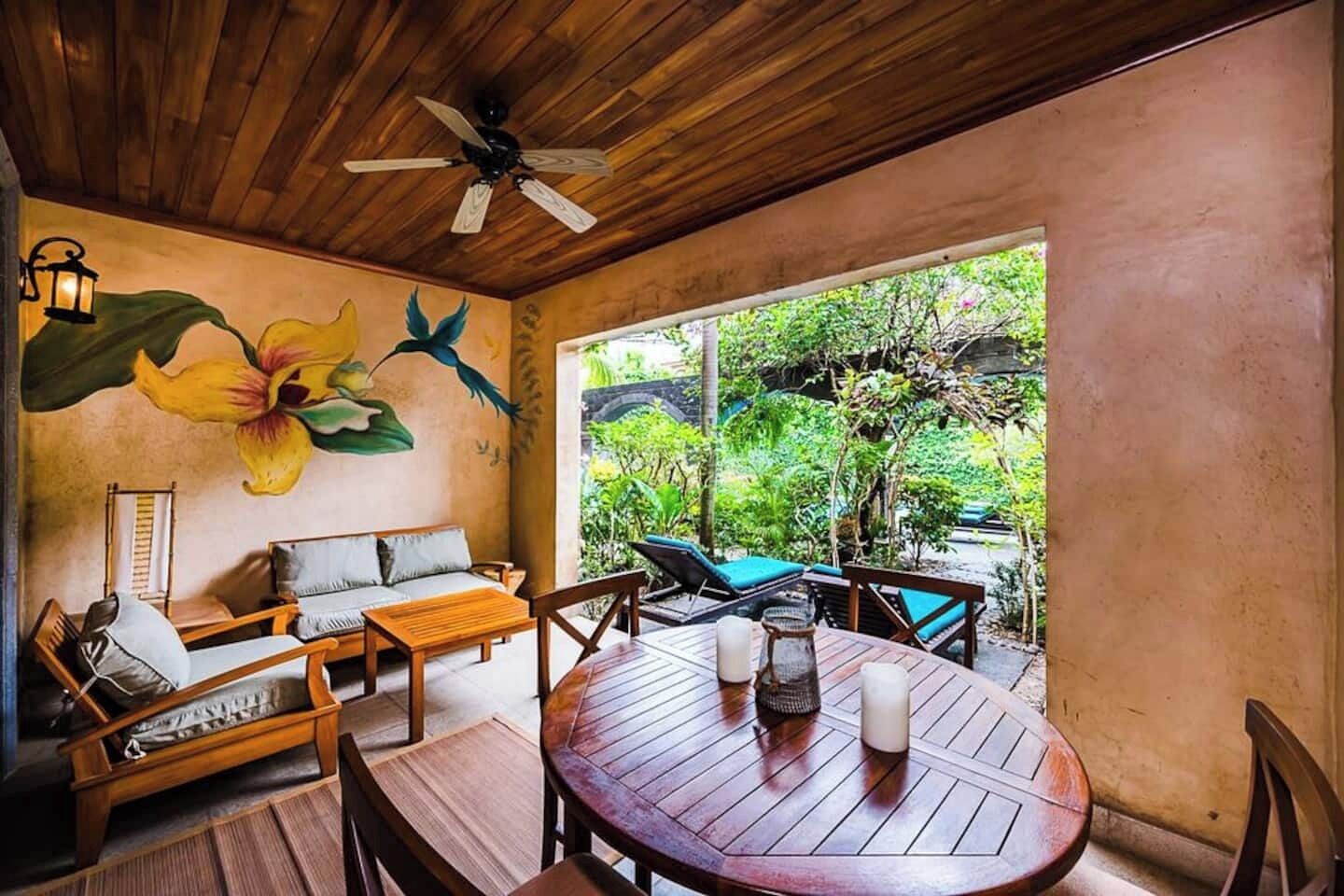 A wooden ceiling fan, a mural of a bird and flower, a round wooden table with candles, and a view of greenery outside.