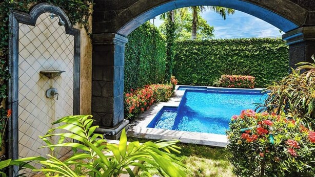 Una piscina con un arco de piedra, rodeada de abundante vegetación y plantas con flores.