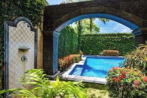A swimming pool with a stone archway, surrounded by lush greenery and flowering plants. - Amanpuri 2, ground Floor unit with kitchen, Grill, Wifi, Sunset view Terrace and pool (Tamarindo)