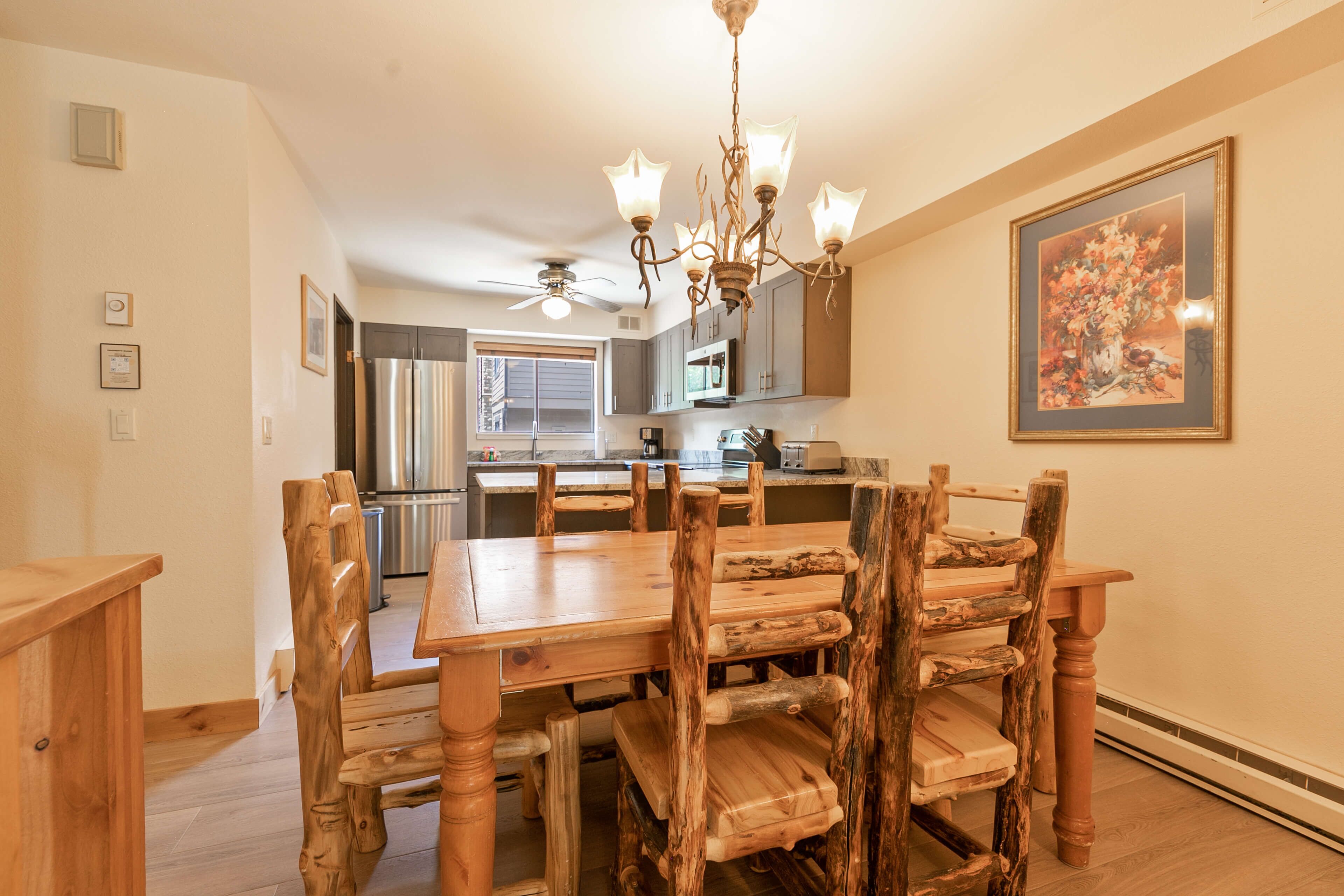 A dining area with a wooden table and chairs, a chandelier, and a kitchen in the background.