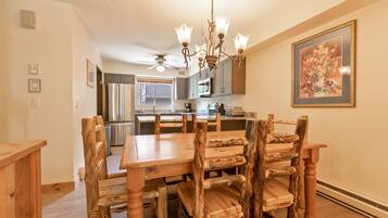 A dining area with a wooden table and chairs, a chandelier, and a kitchen in the background.