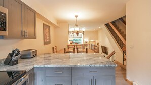 A kitchen with granite countertops, a built-in oven, and a microwave. There is a dining area with a chandelier and wooden chairs.