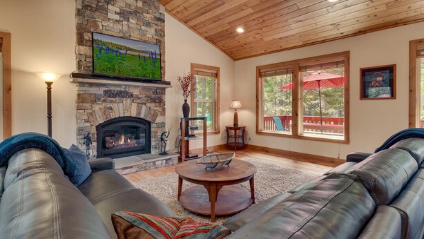 A cozy living room with leather sofas, a stone fireplace, a television mounted on the wall, and large windows with a view of the outdoors.