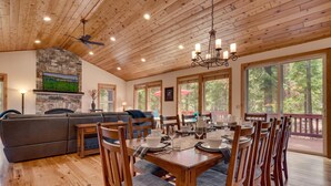A spacious dining area with a wooden table set for four, surrounded by wooden chairs. A stone fireplace with a TV mounted above it is visible in the background.