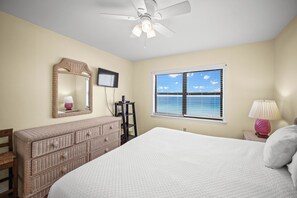Quarto com cama, cômoda, espelho, ventilador de teto, janela com persianas e mesa de cabeceira com abajur.