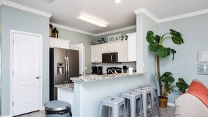 Eine moderne Küche mit weißen Schränken, einer Granit-Arbeitsplatte und Edelstahlgeräten. Es sind drei Barhocker und ein Mülleimer vorhanden.