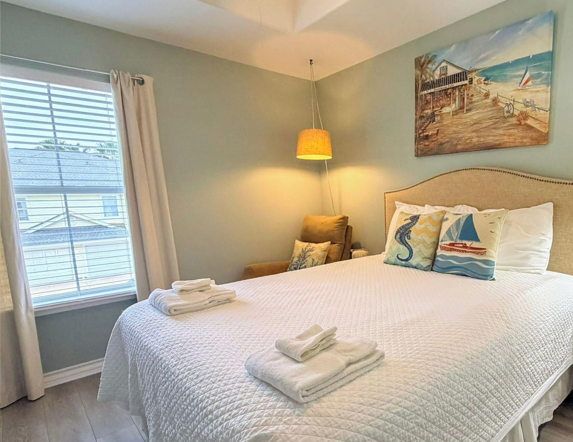 Una cama bien tendida con ropa y toallas blancas, un cuadro de una playa en la pared, y una ventana con cortinas.