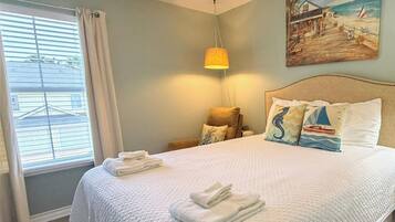 Una cama bien tendida con ropa y toallas blancas, un cuadro de una playa en la pared, y una ventana con cortinas.