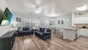A spacious living area with a dark gray sofa set, a dining table with chairs, and a kitchen area with white cabinets and a countertop. - Remodeled Coastal Retreat | Heated Pool, Private Patio & King Beds (Corpus Christi)