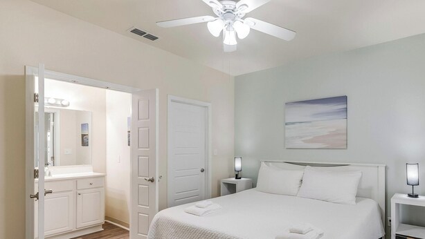 A bedroom with a bed, a ceiling fan, a nightstand with a lamp, a door, and a bathroom with a vanity and mirror. - Remodeled Coastal Retreat | Heated Pool, Private Patio & King Beds (Corpus Christi)