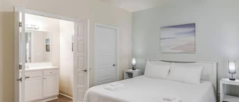 A bedroom with a bed, a ceiling fan, a nightstand with a lamp, a door, and a bathroom with a vanity and mirror.