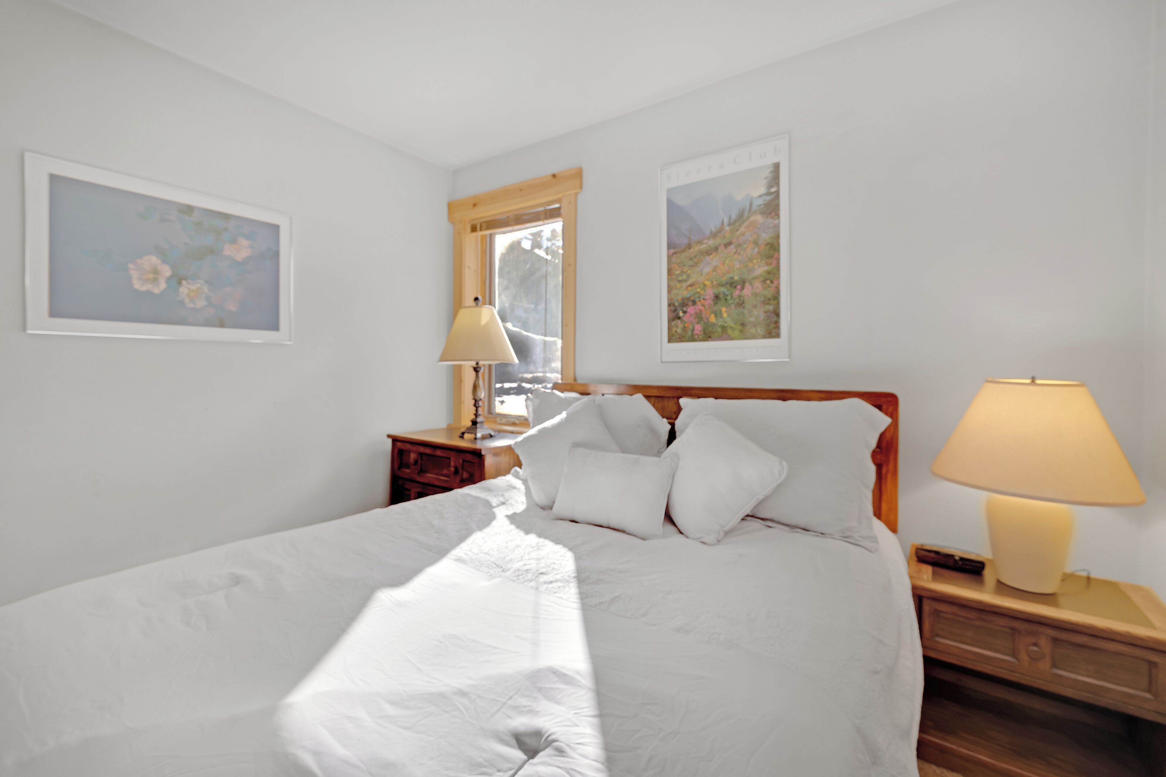 A neatly made bed with white linens, two bedside lamps, a nightstand with a lamp, and two framed pictures on the wall.