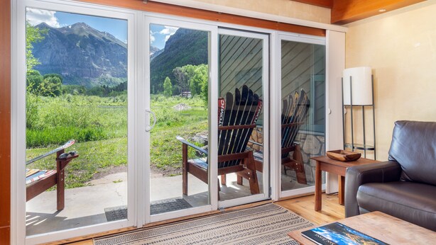 Une pièce dotée d’une grande porte-fenêtre coulissante donnant sur une vue sur les montagnes, d’une chaise en bois, d’un canapé en cuir et d’une petite table sur laquelle se trouve un bol.