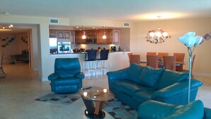 A modern living room with a blue sofa, a glass coffee table, and a kitchen in the background. - Direct Ocean 3 bed, 3 1/2 bath (Cocoa Beach)