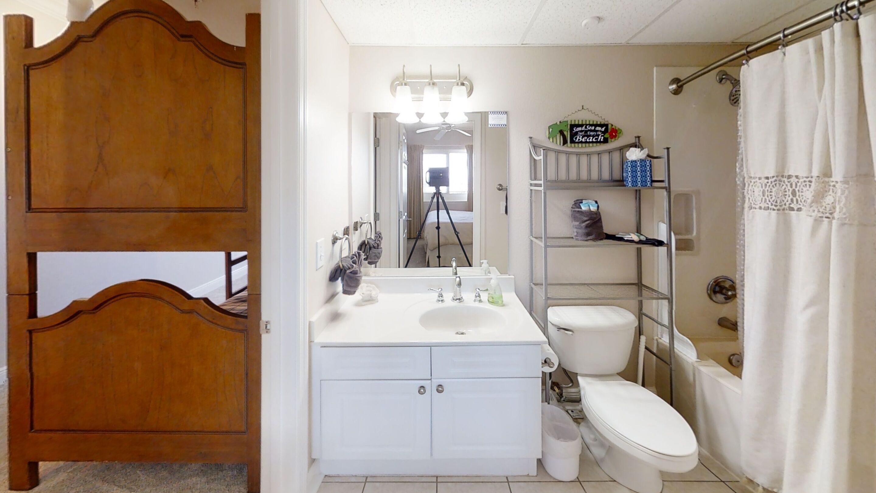 Une salle de bain avec une porte en bois, un lavabo blanc, des toilettes, un rideau de douche et une étagère contenant des articles de toilette.